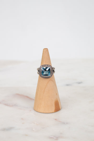 Silver ring with a blue gemstone on a wooden cone against a marble background