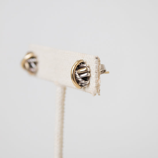 A close-up of a pair of silver-toned textured stud earrings with gold and silver details displayed against a cream-colored display stand and  against a light gray background.