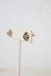 A close-up of a pair of silver-toned textured stud earrings with gold and silver details displayed against a cream-colored display stand and  against a light gray background.
