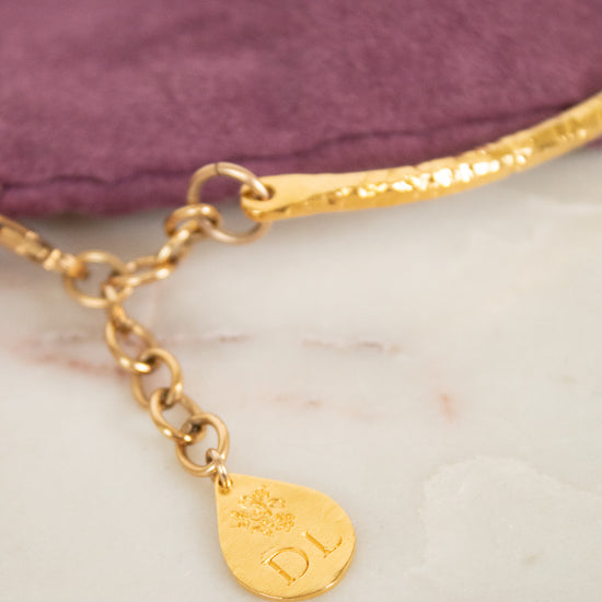 Gold chain bracelet with a textured band, a lobster clasp, and a teardrop-shaped gold tag engraved with 'DL' and a floral motif, resting on a white marble surface next to a purple velvet pouch.