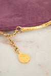 Gold chain bracelet with a textured band, a lobster clasp, and a teardrop-shaped gold tag engraved with 'DL' and a floral motif, resting on a white marble surface next to a purple velvet pouch.