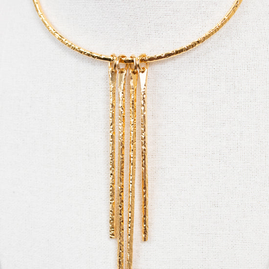 A gold-toned textured necklace displayed on a white fabric mannequin bust. The necklace features a circular top band with five dangling vertical bars hanging from a central connector.