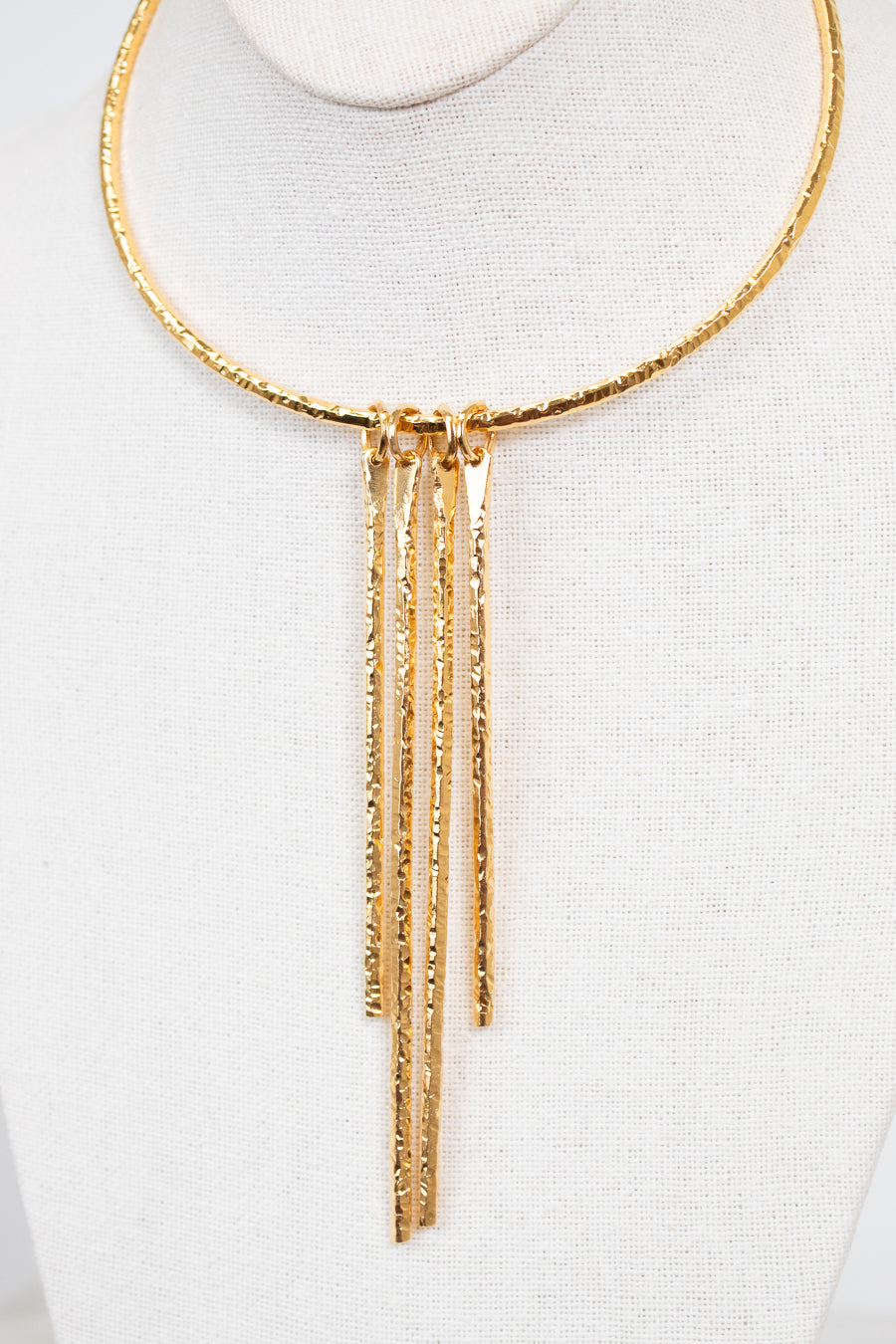A gold-toned textured necklace displayed on a white fabric mannequin bust. The necklace features a circular top band with five dangling vertical bars hanging from a central connector.