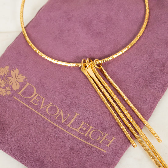 A gold-toned textured bangle necklace with three dangling chain strands, displayed on a purple velvet drawstring pouch printed with the brand name Devon Leigh.