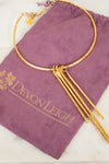 A gold-toned textured bangle necklace with three dangling chain strands, displayed on a purple velvet drawstring pouch printed with the brand name Devon Leigh.
