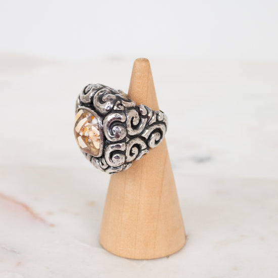 Silver ring with intricate design on a wooden cone against a light background