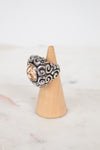 Silver ring with intricate design on a wooden cone against a light background