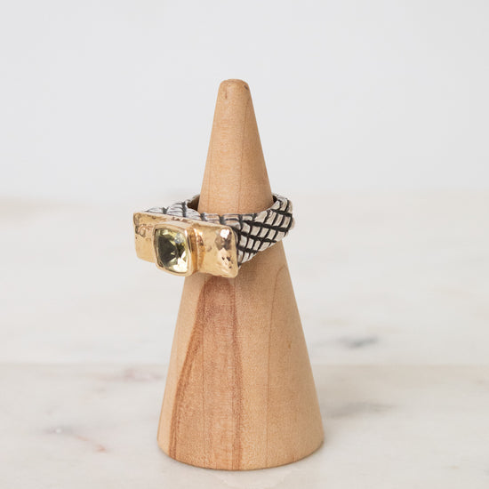 A gold and black patterned ring displayed on a small wooden cone stand against a soft white background.