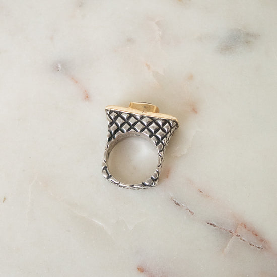 A silver ring with a textured geometric pattern and a gold accent on the top, resting on a light beige marble surface.