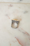 A silver ring with a textured geometric pattern and a gold accent on the top, resting on a light beige marble surface.