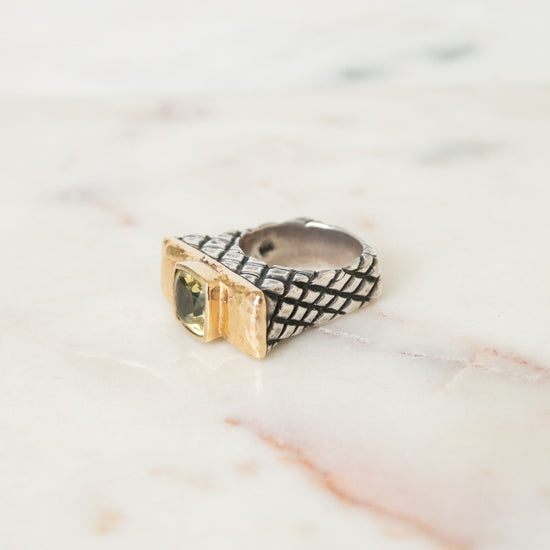A textured ring with a black-and-white woven pattern and a gold-toned bezel-set green stone, resting on a light marble surface.