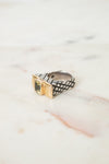 A textured ring with a black-and-white woven pattern and a gold-toned bezel-set green stone, resting on a light marble surface.