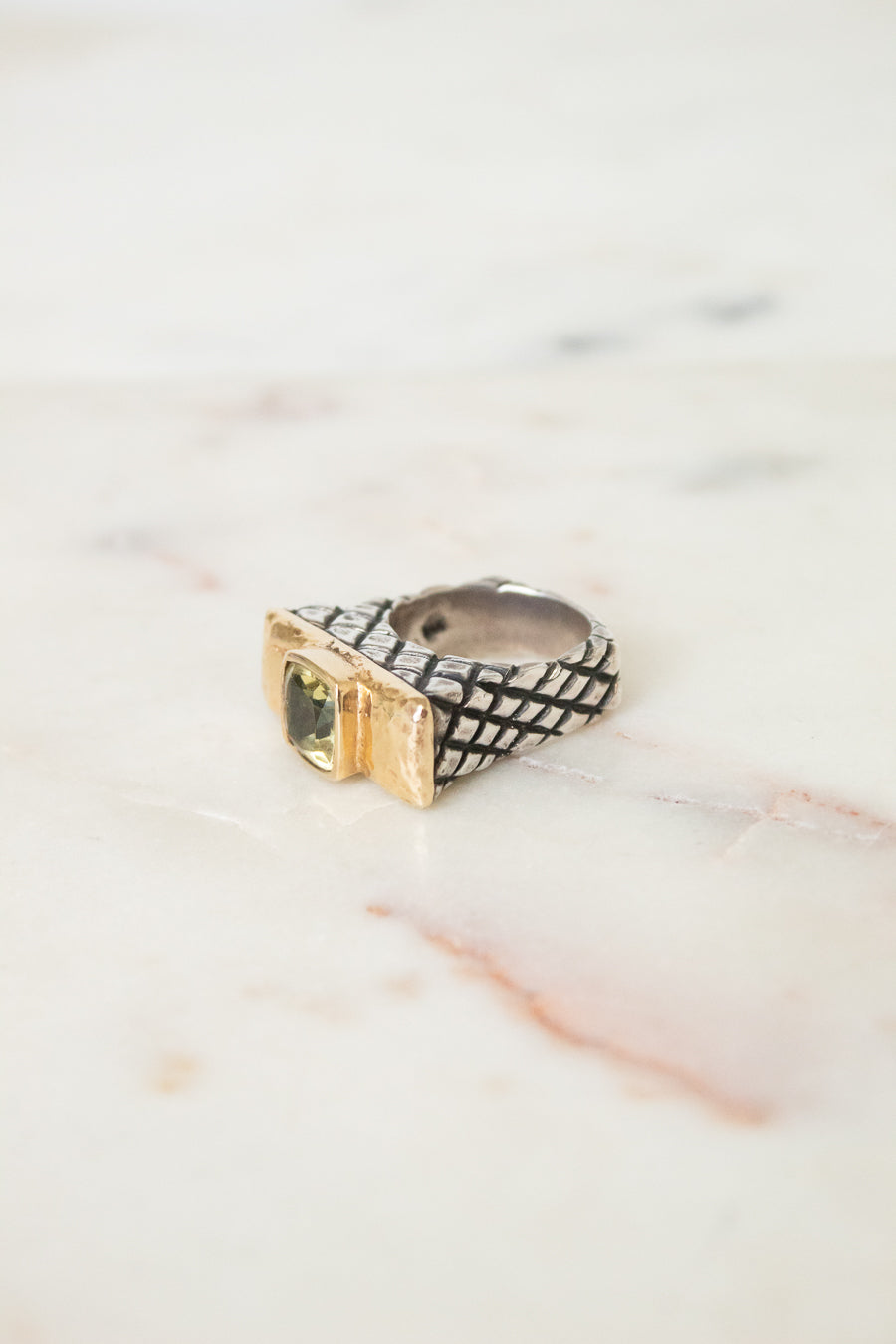 A textured ring with a black-and-white woven pattern and a gold-toned bezel-set green stone, resting on a light marble surface.