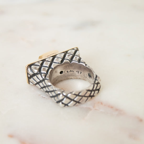 A textured sterling silver ring with a basketweave pattern and a gold-toned accent, resting on a light marble surface.