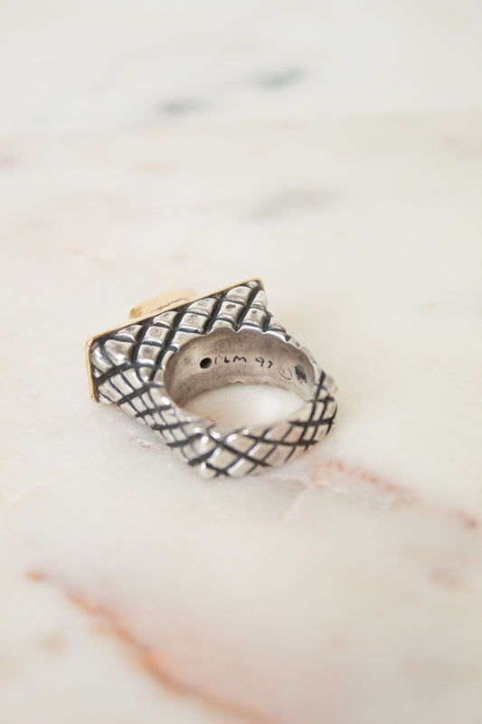 A textured sterling silver ring with a basketweave pattern and a gold-toned accent, resting on a light marble surface.