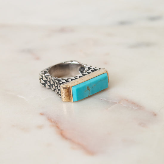A silver ring with a textured band and a rectangular turquoise stone set in gold, resting on a light marble surface.