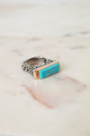 A silver ring with a textured band and a rectangular turquoise stone set in gold, resting on a light marble surface.
