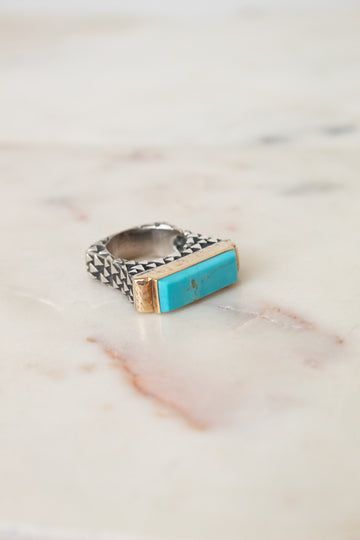 A silver ring with a textured band and a rectangular turquoise stone set in gold, resting on a light marble surface.