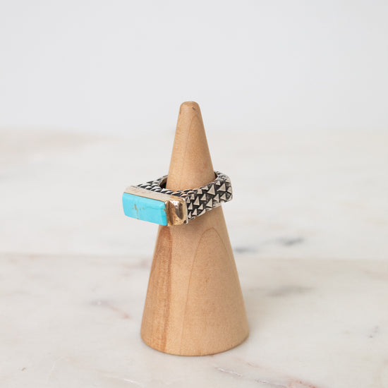 A silver ring with a rectangular turquoise stone, displayed on a light wood cone stand against a white marble surface and white background.