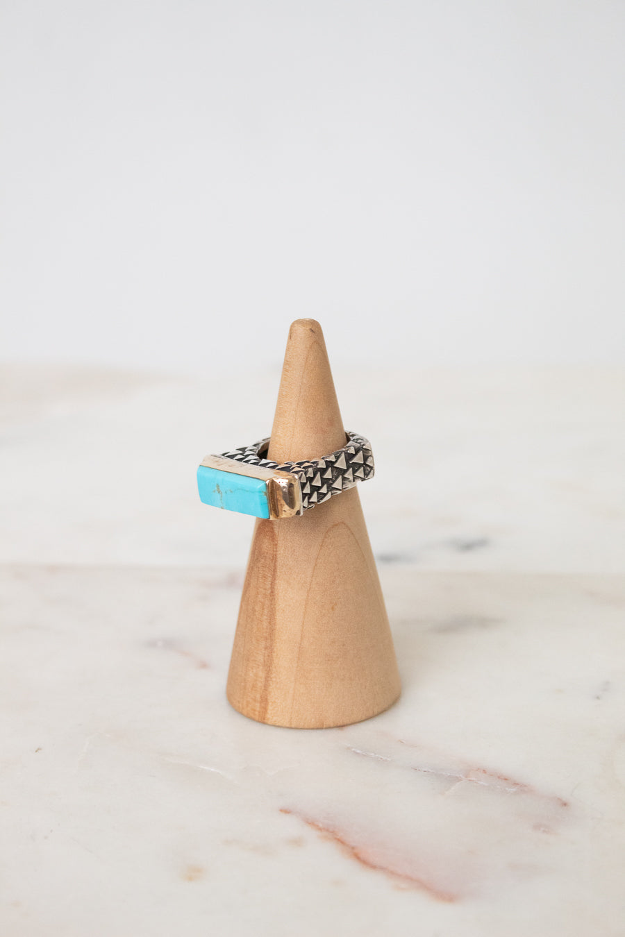 A silver ring with a rectangular turquoise stone, displayed on a light wood cone stand against a white marble surface and white background.