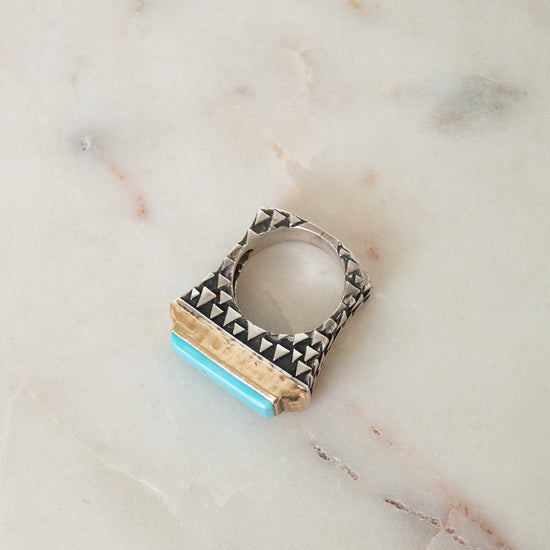 A geometric silver ring with black triangular inlay and a turquoise blue accent, resting on a light beige marble surface.