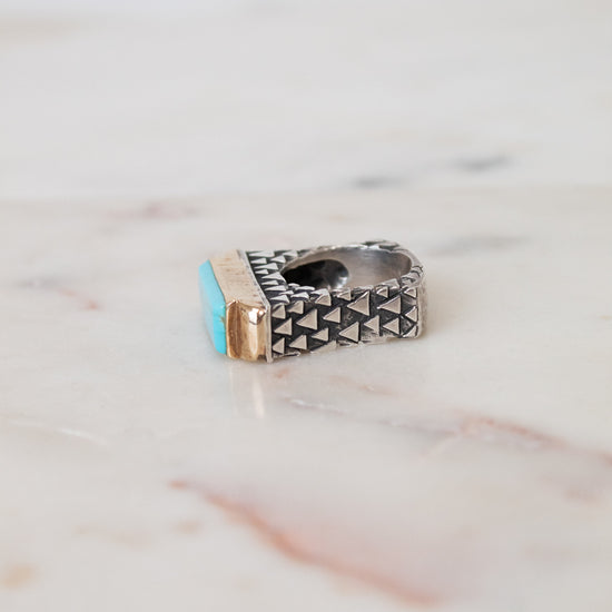A silver-toned geometric ring with a small turquoise blue stone and a gold accent, resting on a light marble surface.