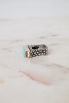 A silver-toned geometric ring with a small turquoise blue stone and a gold accent, resting on a light marble surface.