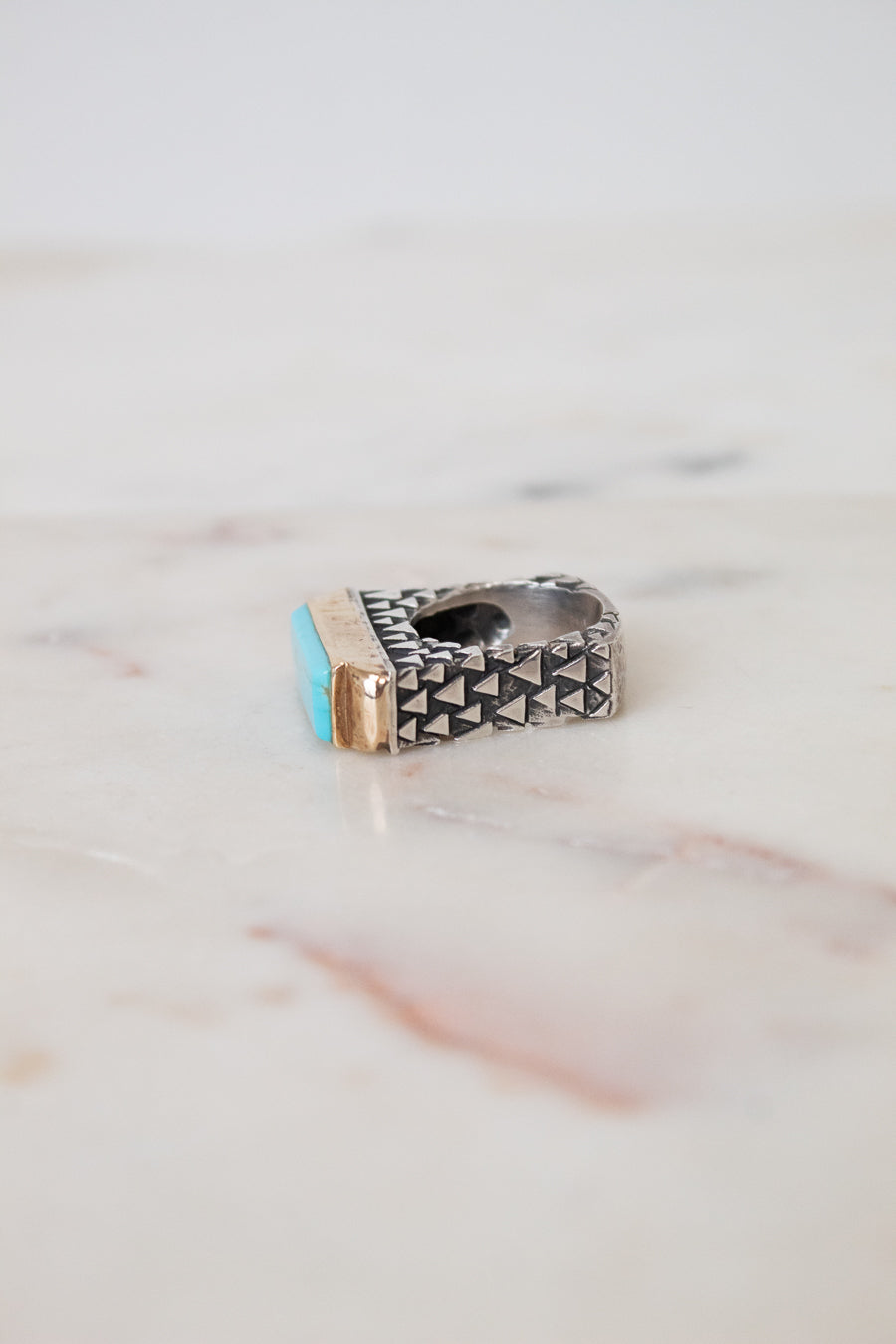 A silver-toned geometric ring with a small turquoise blue stone and a gold accent, resting on a light marble surface.
