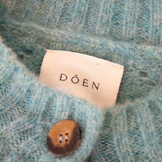 Close-up of a teal knitted garment with a 'Doen' label and button.