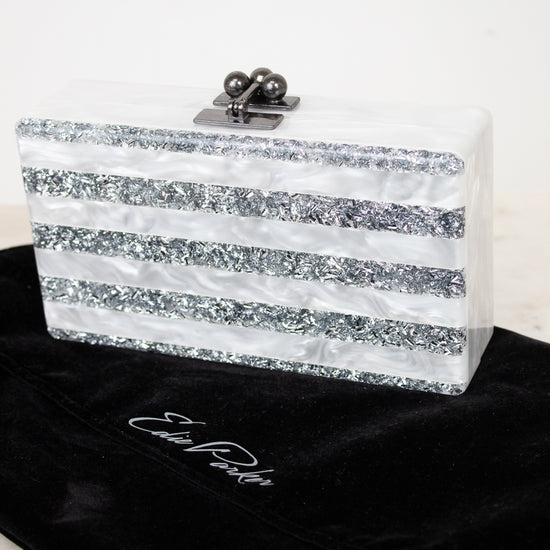 A rectangular clutch bag with horizontal silver glitter stripes and a silver clasp, displayed on a black velvet pouch with  EDIE PARKER  embroidered on it against a white background.