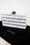 A rectangular clutch bag with horizontal silver glitter stripes and a silver clasp, displayed on a black velvet pouch with  EDIE PARKER  embroidered on it against a white background.