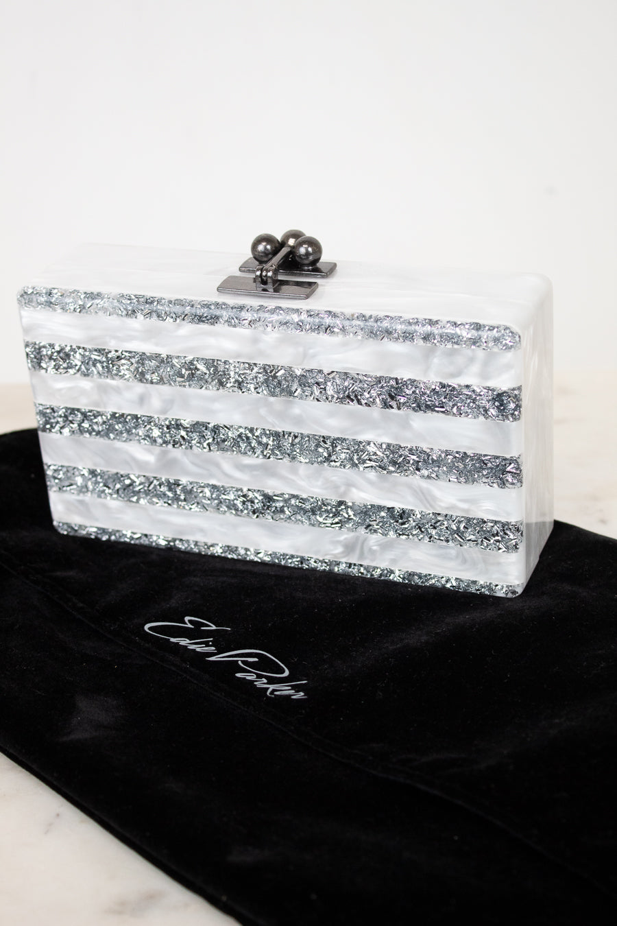 A rectangular clutch bag with horizontal silver glitter stripes and a silver clasp, displayed on a black velvet pouch with  EDIE PARKER  embroidered on it against a white background.