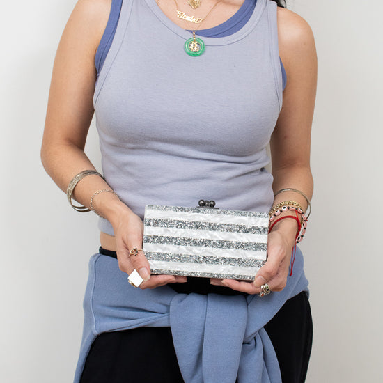 A woman wearing a light blue ribbed tank top with darker blue trim, holding a silver glitter clutch bag. She is also wearing a light blue sweatshirt tied around her waist and black pants, set against a plain light gray background.