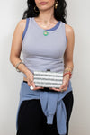 A woman wearing a light blue ribbed tank top with darker blue trim, holding a silver glitter clutch bag. She is also wearing a light blue sweatshirt tied around her waist and black pants, set against a plain light gray background.