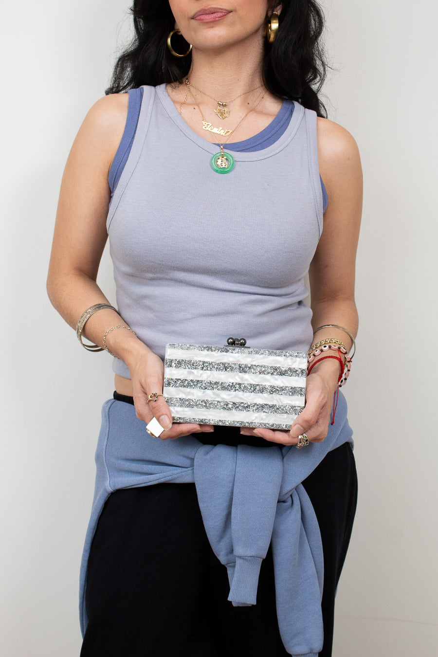 A woman wearing a light blue ribbed tank top with darker blue trim, holding a silver glitter clutch bag. She is also wearing a light blue sweatshirt tied around her waist and black pants, set against a plain light gray background.