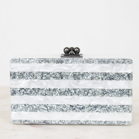 A rectangular clutch bag with horizontal stripes of silver glitter and white faux mother-of-pearl, featuring a black metal clasp with three dark beads on top, set against a white background.