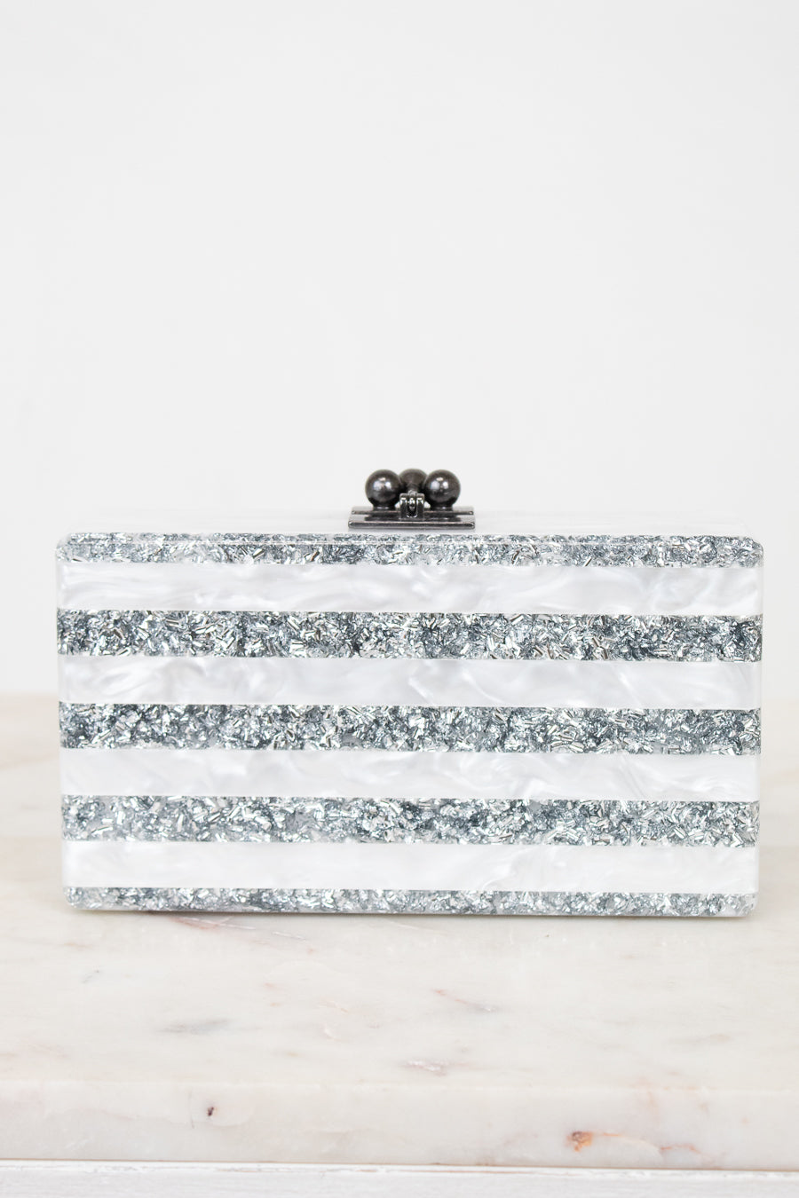 A rectangular clutch bag with horizontal stripes of silver glitter and white faux mother-of-pearl, featuring a black metal clasp with three dark beads on top, set against a white background.