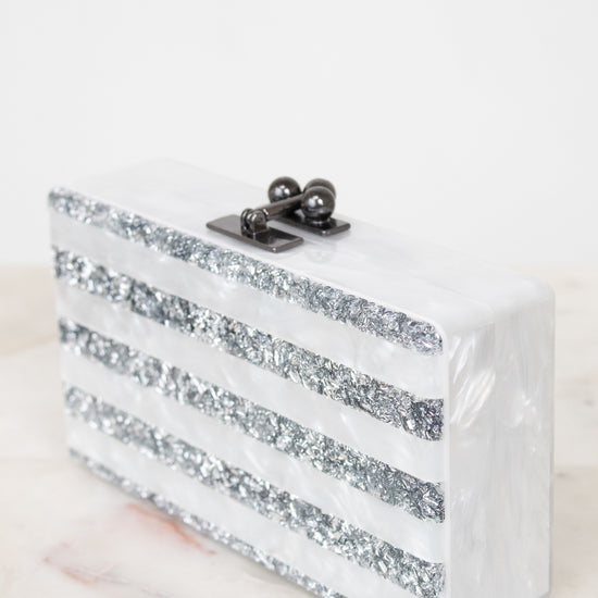 A rectangular white resin clutch bag with horizontal silver glitter stripes and a black metal clasp, resting on a light marble surface against a white background.