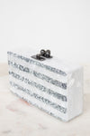 A rectangular white resin clutch bag with horizontal silver glitter stripes and a black metal clasp, resting on a light marble surface against a white background.
