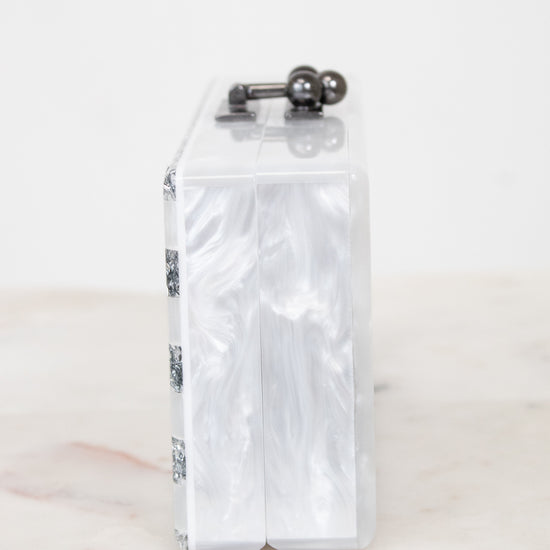 A slim rectangular clutch bag made of glossy white marbled faux mother-of-pearl, featuring a dark metal top handle with two spherical accents, resting on a light marble surface against a white background.