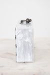 A slim rectangular clutch bag made of glossy white marbled faux mother-of-pearl, featuring a dark metal top handle with two spherical accents, resting on a light marble surface against a white background.
