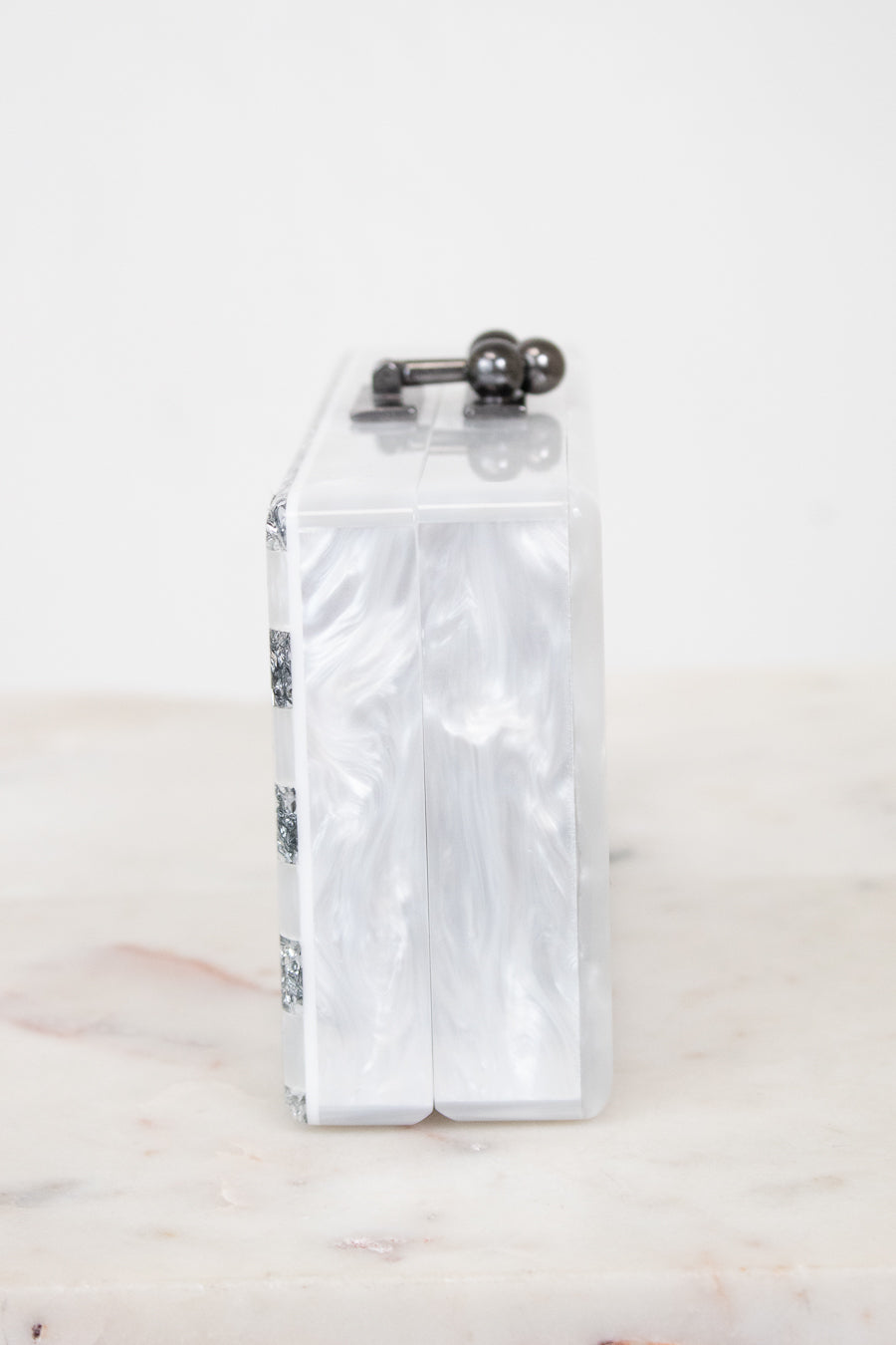 A slim rectangular clutch bag made of glossy white marbled faux mother-of-pearl, featuring a dark metal top handle with two spherical accents, resting on a light marble surface against a white background.