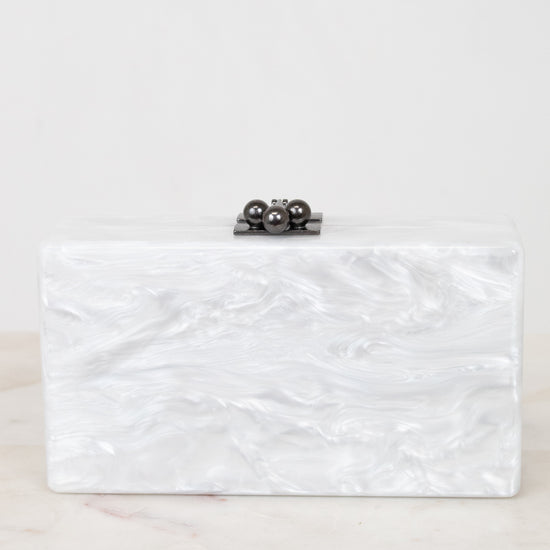 A rectangular clutch bag with a white and silver marbled shell-like finish, featuring a small black metal clasp with three dark pearls on top, resting on a light marble surface against a white background.