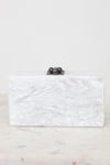 A rectangular clutch bag with a white and silver marbled shell-like finish, featuring a small black metal clasp with three dark pearls on top, resting on a light marble surface against a white background.