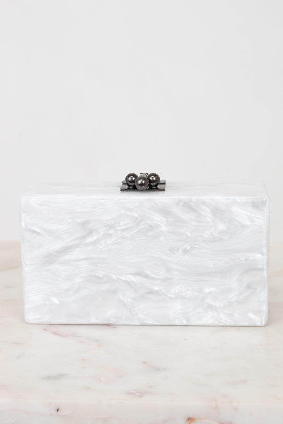 A rectangular clutch bag with a white and silver marbled shell-like finish, featuring a small black metal clasp with three dark pearls on top, resting on a light marble surface against a white background.