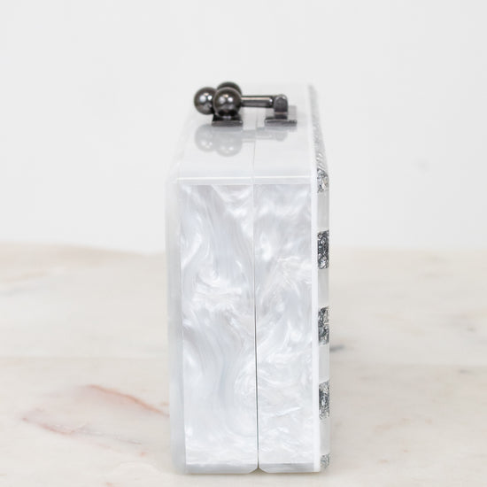 A rectangular clutch bag with a glossy white and silver marbled shell-like pattern, featuring a dark metal top clasp and a side chain detail, displayed on a light marble surface against a white background.