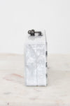 A rectangular clutch bag with a glossy white and silver marbled shell-like pattern, featuring a dark metal top clasp and a side chain detail, displayed on a light marble surface against a white background.