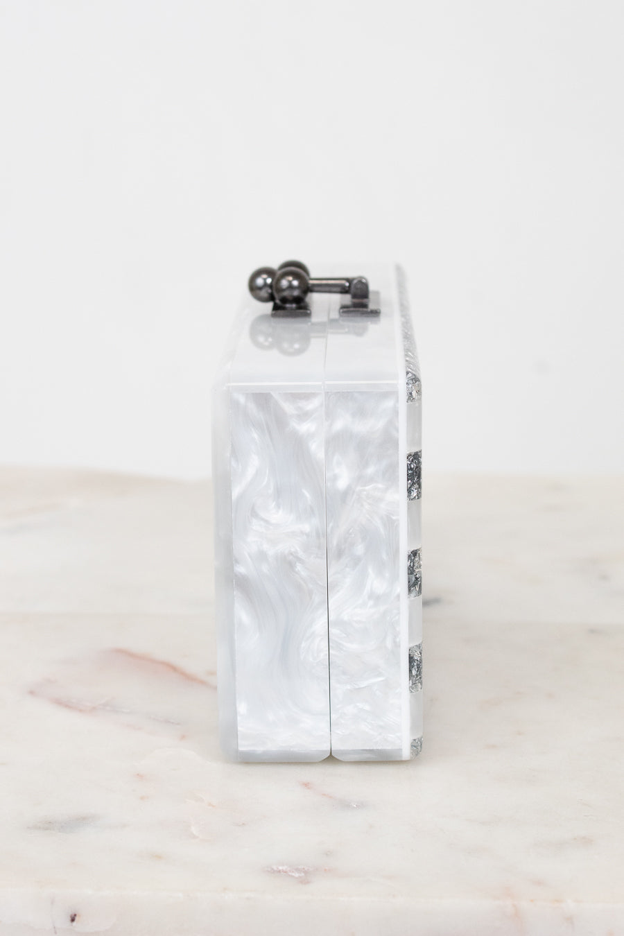 A rectangular clutch bag with a glossy white and silver marbled shell-like pattern, featuring a dark metal top clasp and a side chain detail, displayed on a light marble surface against a white background.