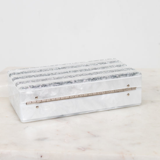 A rectangular white resin jewelry box with horizontal textured silver bands on the lid and a small gold-tone metal bar on the front, displayed on a white marble surface against a white background.