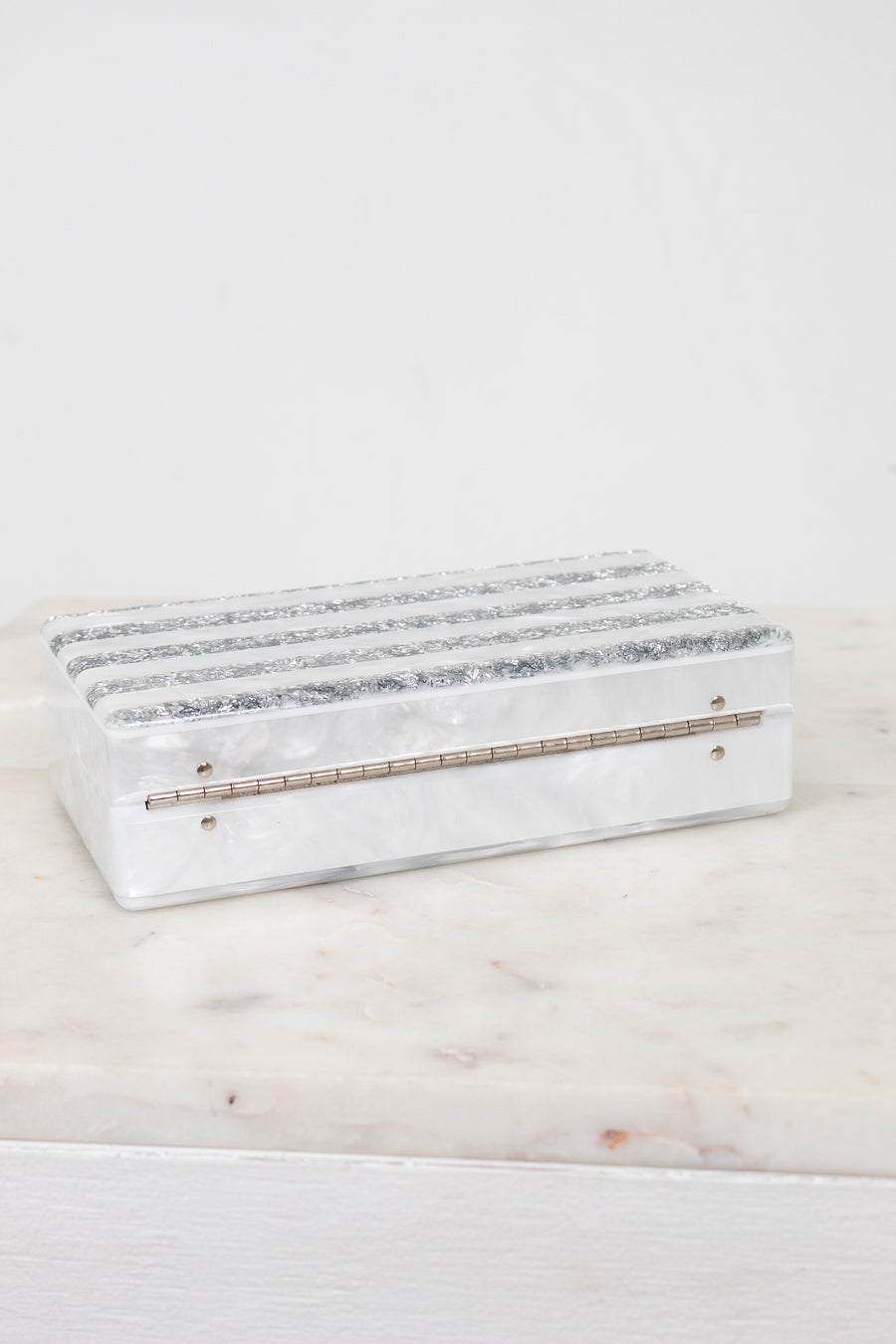 A rectangular white resin jewelry box with horizontal textured silver bands on the lid and a small gold-tone metal bar on the front, displayed on a white marble surface against a white background.